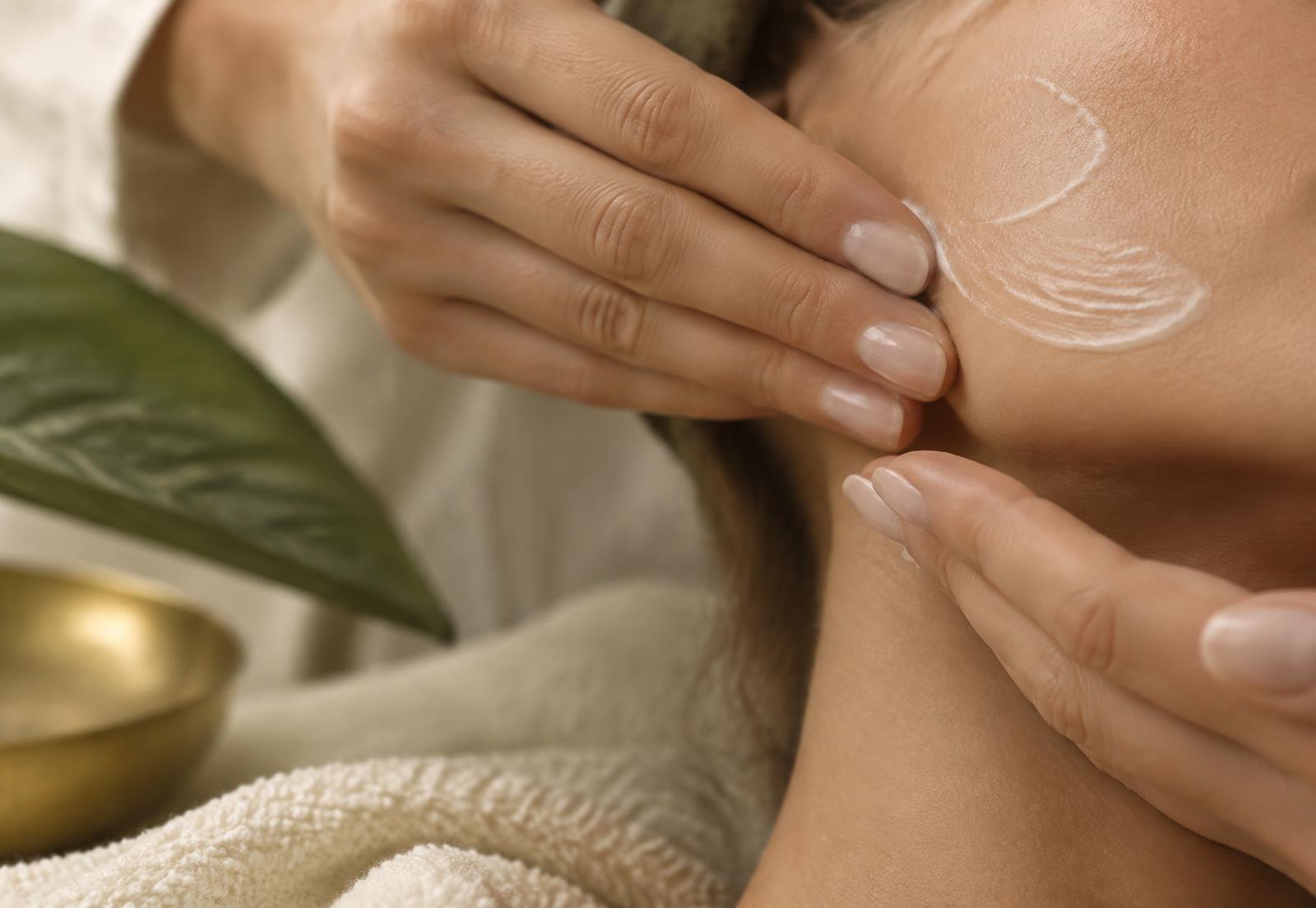 Gros plan de mains appliquant un soin hydratant sur la joue, visage hors cadre